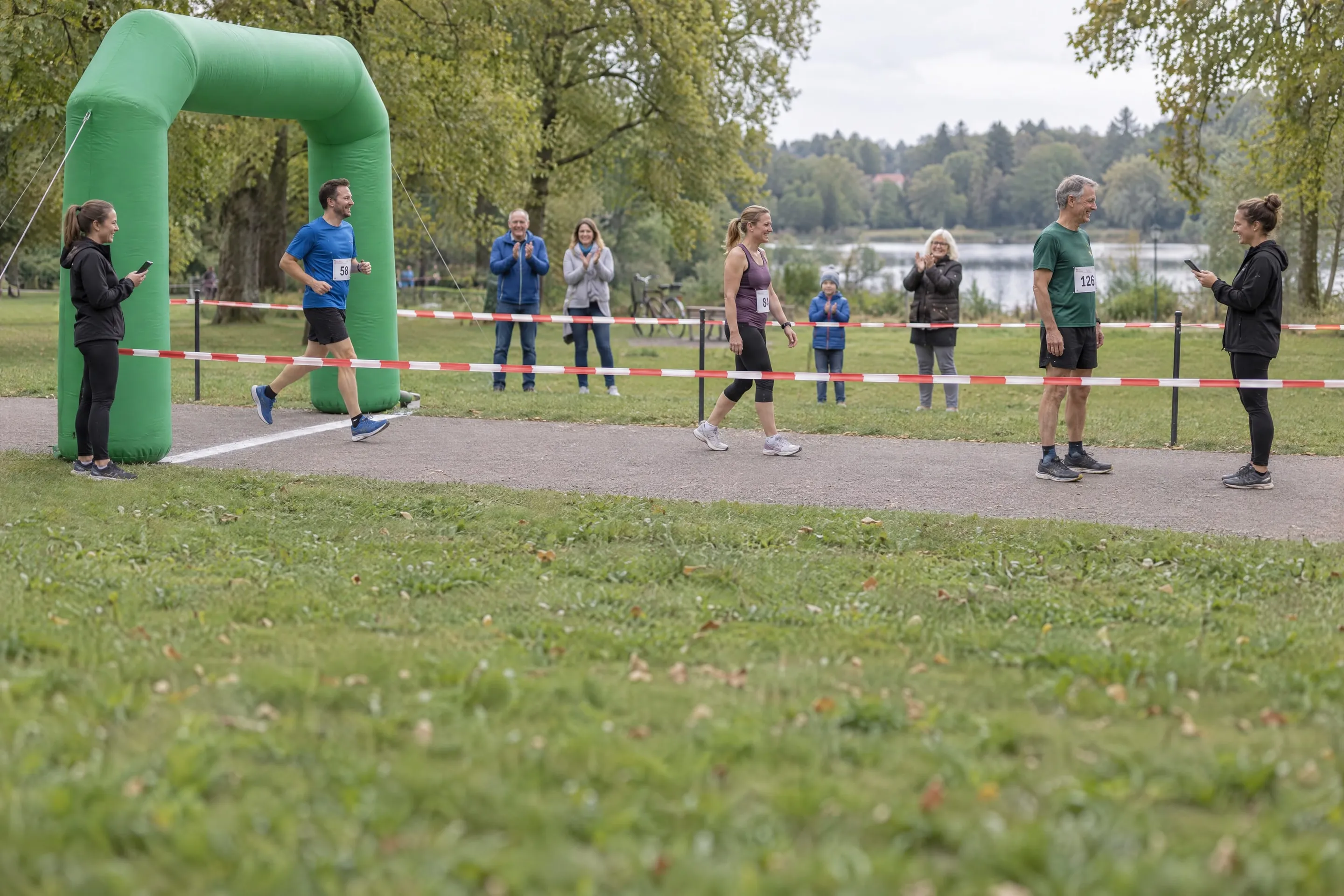Ein Läufer kommt unter einem grünen Zielbogen ins Ziel, daneben stoppt eine Helferin die Zeit per Smartphone; im abgesperrten Zielkanal gehen die Finisher hintereinander zu einer Helferin, die ihre Startnummern erfasst, Zuschauer applaudieren