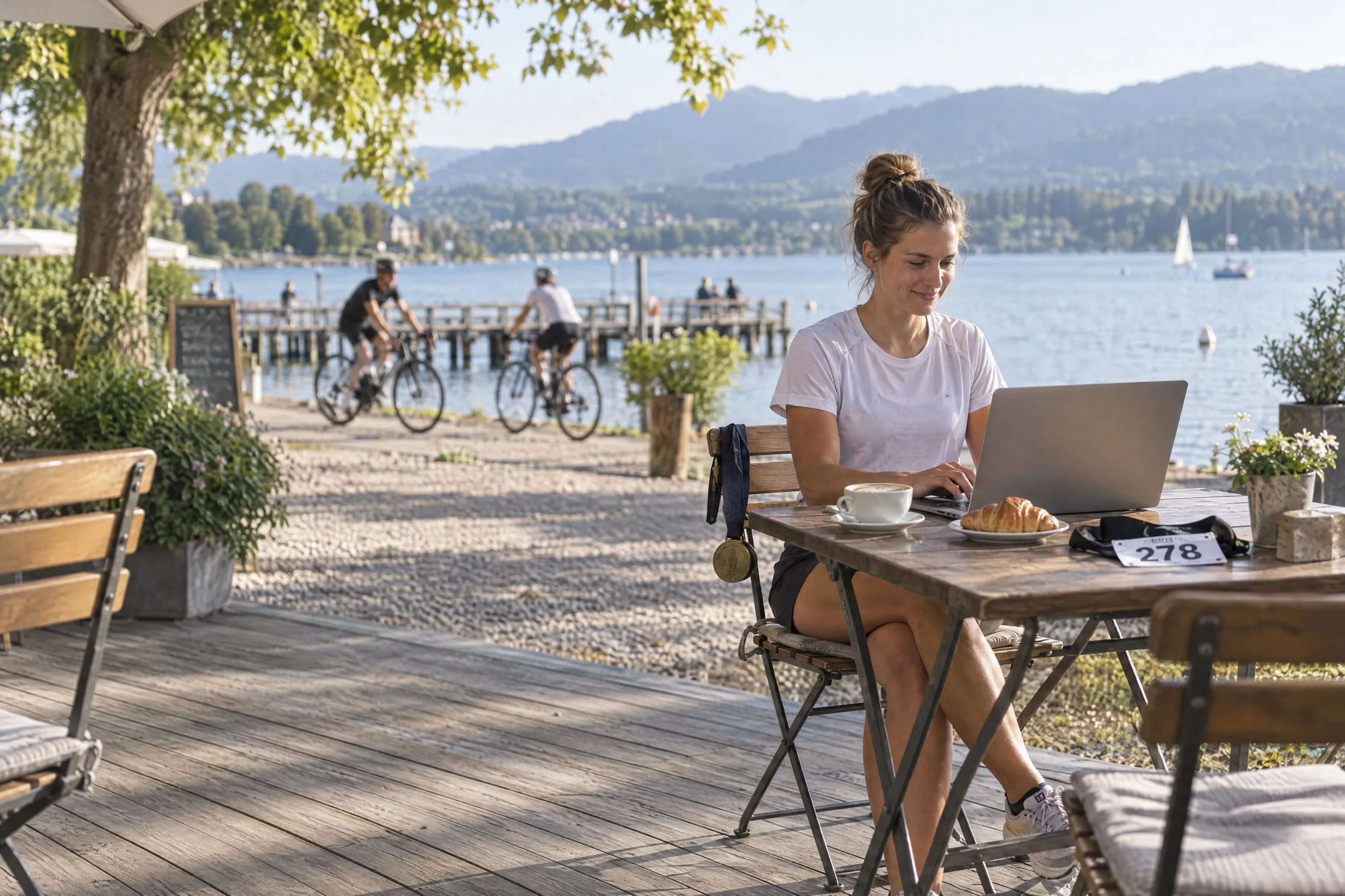 Junge Triathletin schreibt am Morgen nach ihrem ersten Langdistanz-Triathlon auf der Terrasse eines Seecafés am Laptop ihren Wettkampfbericht, die Finisher-Medaille hängt am Stuhl